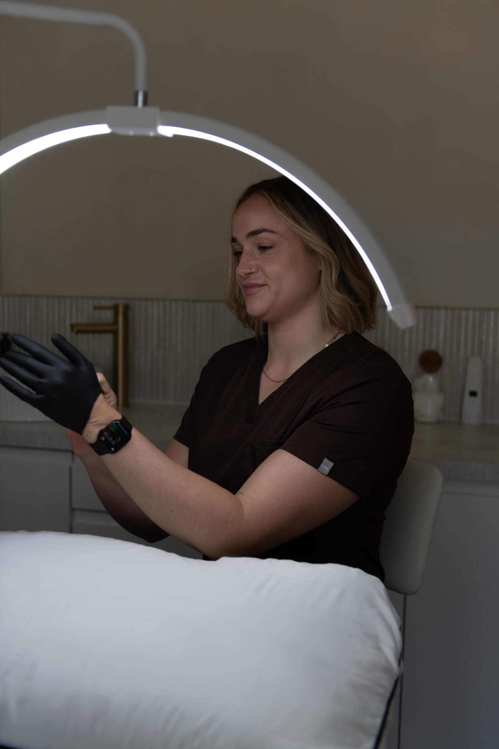 Chloe in her treatment room, putting on black gloves beside the treatment couch under an LED lamp
