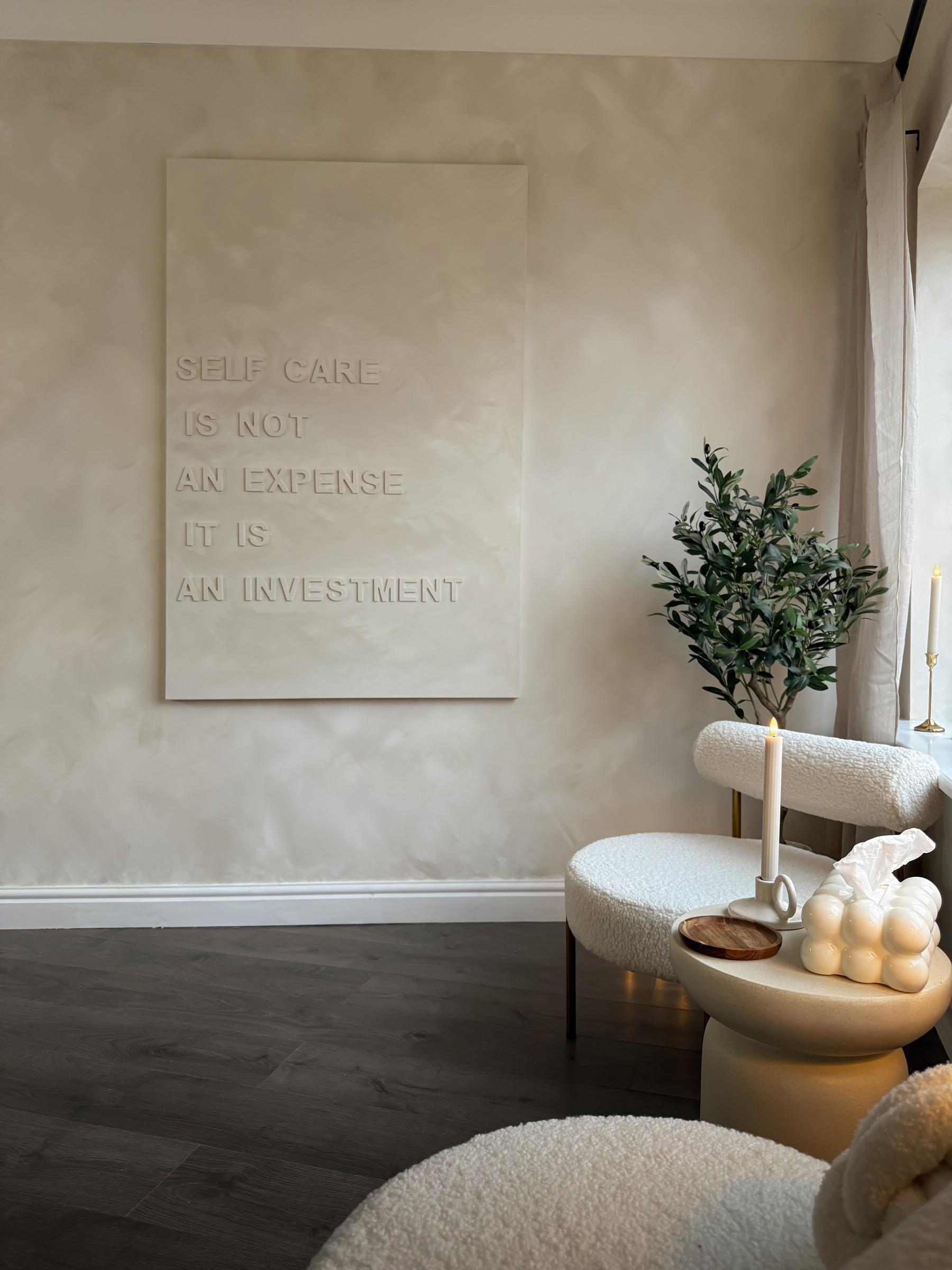 The calm waiting area at The Skin Lab: boucle chairs, an olive tree, a candle and a wall piece reading ‘self care is not an expense, it is an investment’